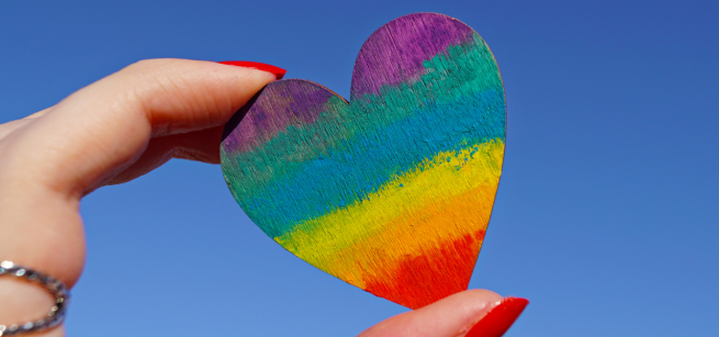 A pale-skinned hand holds a rainbow-painted heart against the sky