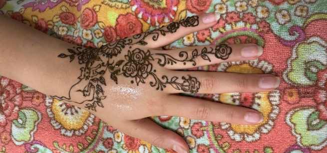 A photo of a hand and partial image of an arm, on a colorful cloth background, featuring a detailed henna design on the skin.