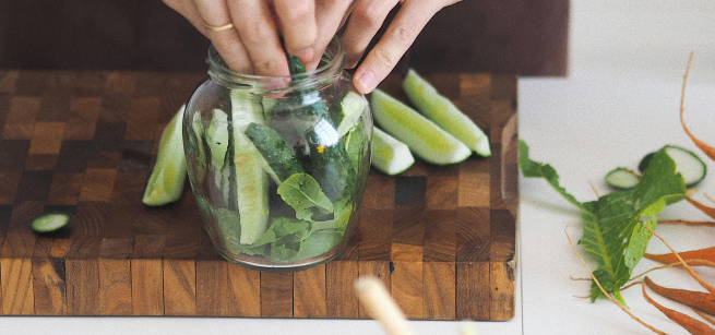Hands putting sliced cucumbers in a glass jar to be pickled. Other veggies in foreground.  