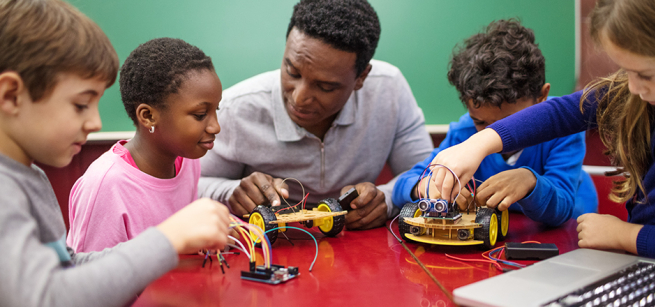 kids sit around a table with an adult examining small electronics