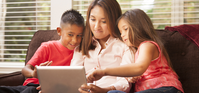 A mother sits on a couch between two children of elementary-school age. They are looking at a computer tablet.