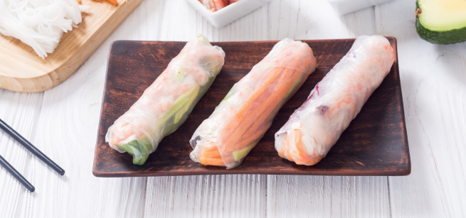 Three fresh spring rolls filled with vegetables are arranged on a rectangular dark plate, with chopsticks placed nearby on a white wooden surface.