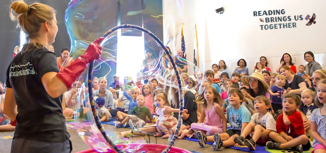 Adult holding a hula hoop, making bubbles, in front of a crowd of children