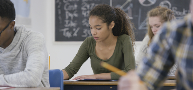 Girl in classroom studying at desk with chalkboard behind her