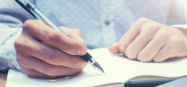Close-up of two hands resting on an open journal, with the right hand holding a pen to write.