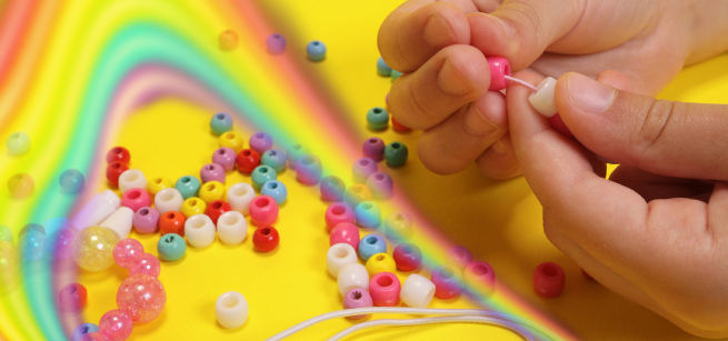 photo of two hands stringing beads on a thread; colorful beads and a rainbow cover the surface below