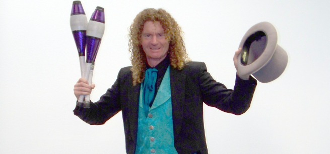 A smiling red haired man holds a top hat and a pair of juggling clubs