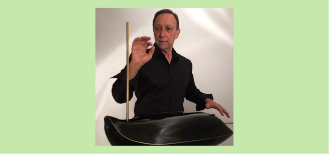 Ed Sussman is wearing a black collared shirt, standing behind a theremin with his hand raised next to it.