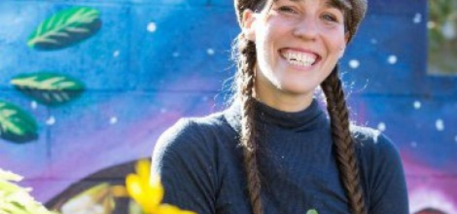 A woman smiling with braided hair in front of a vibrant colorful mural. 