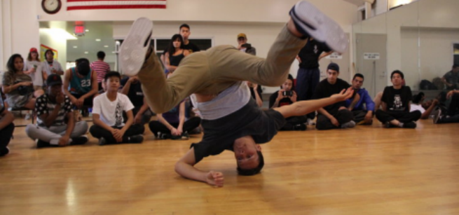 Breakdance with head on the ground and people looking on in a gym