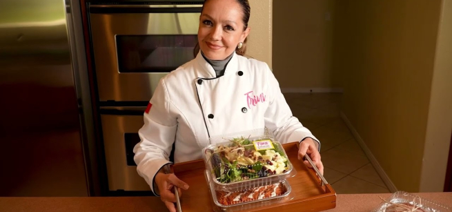 Women wearing white chef's coat in home kitchen holding up boxed meals on a tray.