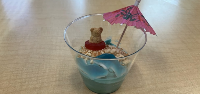 A cup of blue pudding on a table decorated with cookie crumbs, candy and a paper umbrella