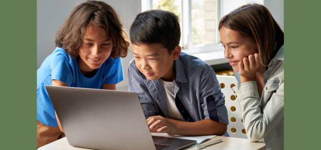 Three kids all looking at a computer smiling. Two boys and a girl who is holding her head in her hand on the tabletop
