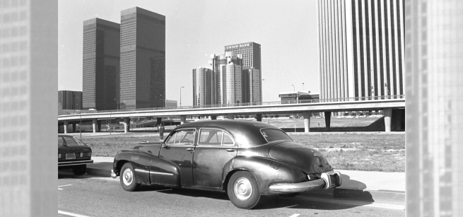 1940s automobile parked on Hope Street in 1970s with LA skyline in the background. 