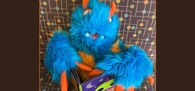 Blue monster puppet with orange claws reading a picture book