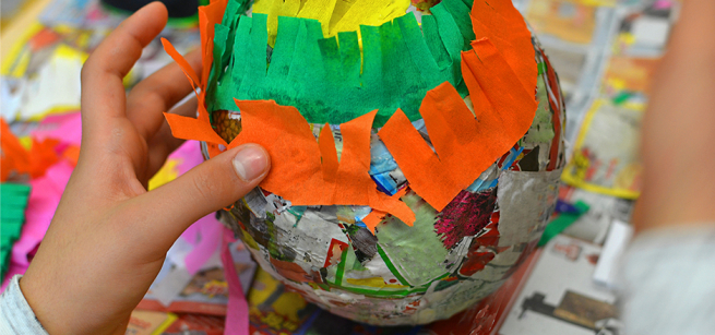 Close-up of hands crafting a colorful object with vibrant orange, green, and yellow paper strips. The scene conveys creativity and focus.