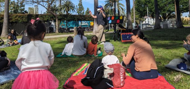 toddlers and adult caregivers seated on a grassy lawn watch a librarian leading a storytime in a park