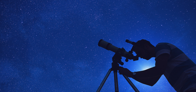 backlit man looks through telescope at night sky