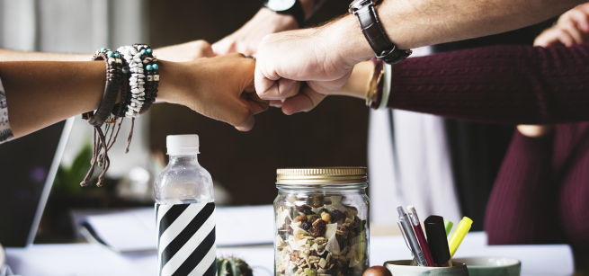 four arms with watches fist bump, meeting in the middle and a water bottle, jar of coins along with notebooks located