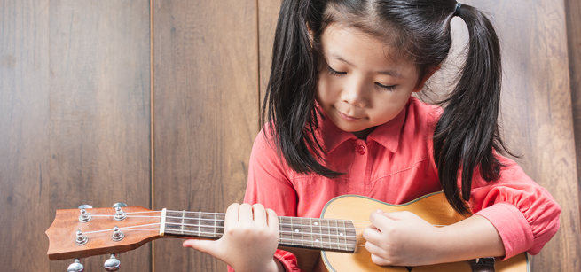 Child with pigtails and a red shirt plays a ukulele.