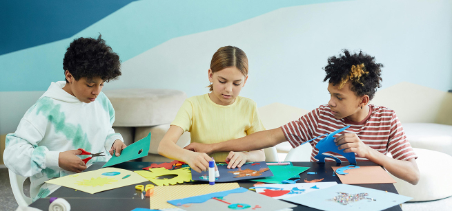 Three children concentrate on making handmade crafts with an assortment of craft supplies.