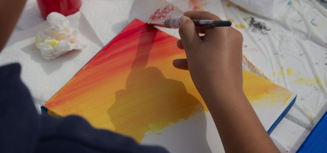 Shows a hand holding a paint brush and a small canvas being painted with orange and yellow paint diagonally from the top left corner on a surface covered with white cloth and a crumpled up paper towel.