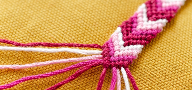 A friendship bracelet made of red, pink, and white string weaved with a chevron pattern against a golden yellow fabric