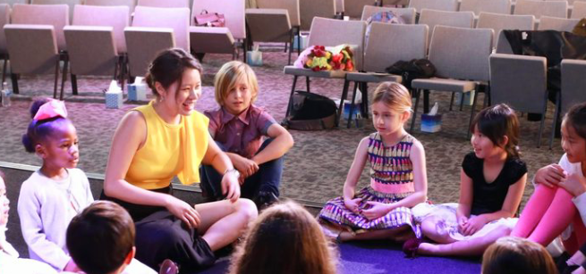 Miss Sarah sitting in a circle with other children, holding her hands up mid-clap