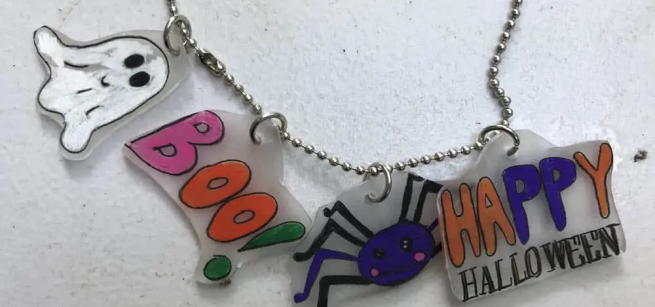 Halloween-themed necklace with charms: a ghost, "BOO!" text, a purple spider, and "HAPPY HALLOWEEN" in colorful, playful fonts.