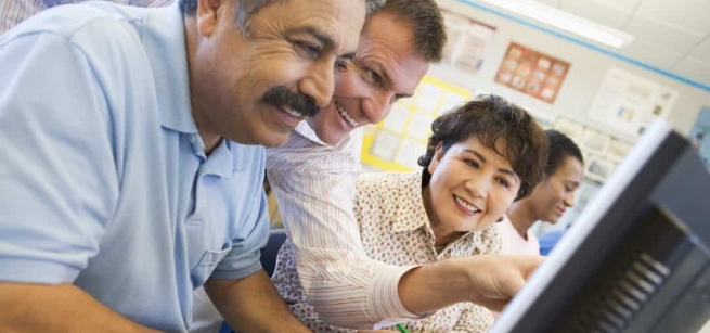 Two adult students looking at a computer screen while receiving instruction from a teacher
