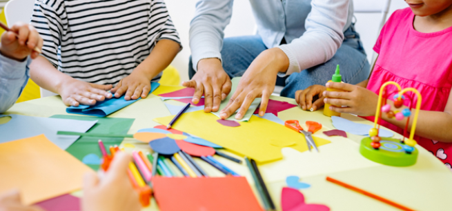 Hands of children and adults are shown as they create art from colorful supplies strewn across a table. 