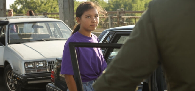 Beans played by Kiawentiio stands on a bridge by a car facing a man in uniform
