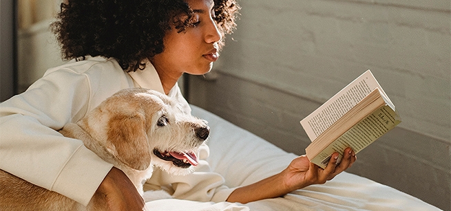 A woman reads a book on a bed with her arm around a dog