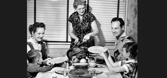 A woman carving a turkey at a table surrounding by two adults and two children
