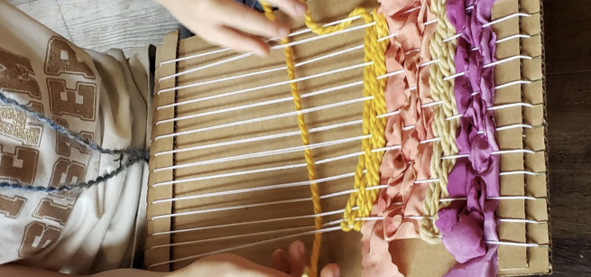 hands weaving yarns and ribbons on a cardboard loom