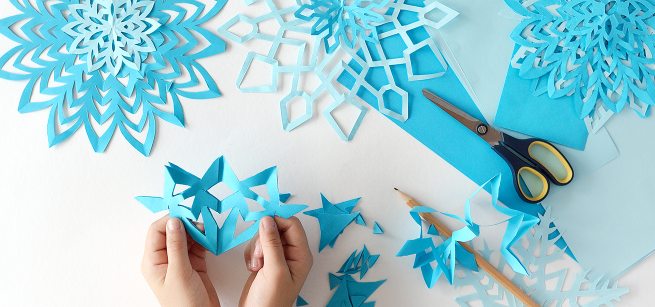 Two hands holding blue paper shaped like a snowflake, various hues of blue craft paper in the background, scissors and a pencil also in the background.