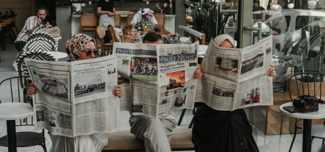 Three ladies sitting in chairs in a row holding up newspapers in front of their faces