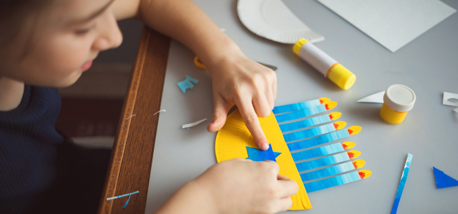 A child decorating a Hanukkah menorah cut-out in shades of blue with nine candles lit.