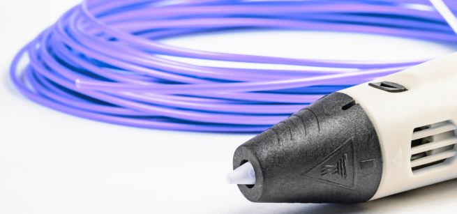 A partial view of a white 3D pen with a black tip angled at the bottom right corner and a spool of purple plasic filament behind it on a white surface.