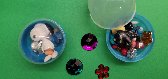 Picture of two capsule containers, one open and one sealed, with various beads and other colorful items in them.