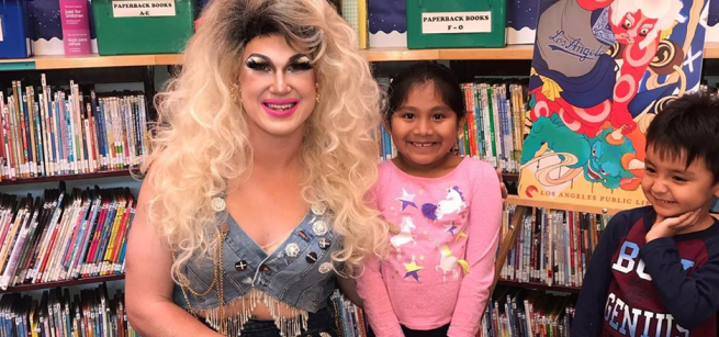 A drag performer in a blonde wig sits in a library with two smiling children.