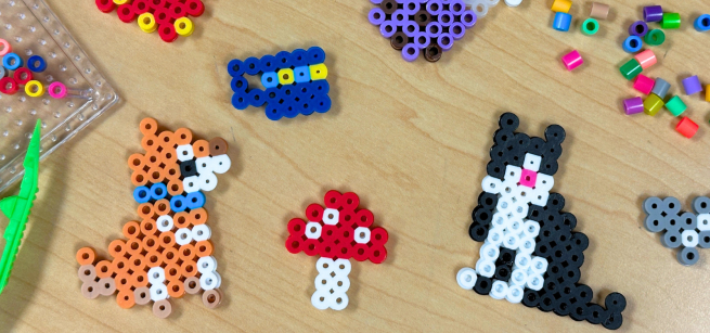Various fuse bead creations such as a mushroom and cat are displayed with fuse bead crafting tools and unused beads.