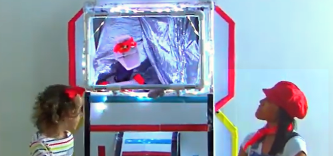 A robot in a red mask and inside a rocket ship speaking to Cheri Moon while a little girl looks at them.