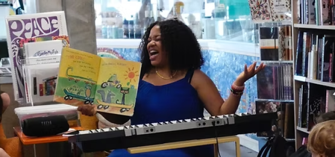 A musician belts out a song while reading a storybook to children. She sits at a musical keyboard and is surrounded by books.