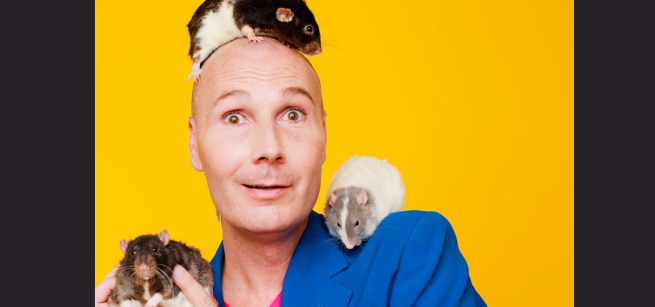 Christopher T. Magician in a blue coat against a yellow background holding guinea pigs.