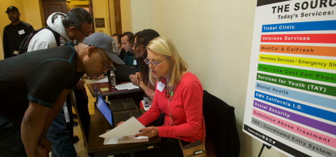 Image of a seated woman in a red sweater at a table helping a standing man with paperwork.