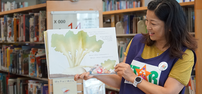 STAR Reader wearing a purple vest holds a full page spread of a picture book in surrounded by the children's library stacks.