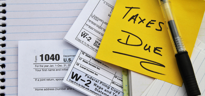 a yellow post it note with Taxes Due and three tax forms on top of a notebook