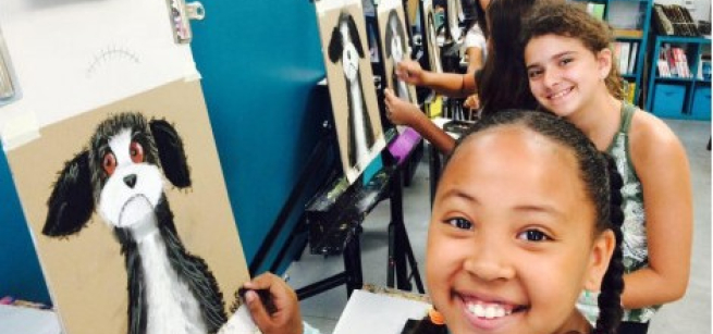 Smiling children in the art studio, seated at their easels with their completed paintings of dog portraits.