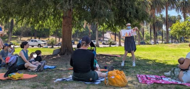 librarian reads at a park in front of toddlers and adult caregivers seated on a grassy lawn